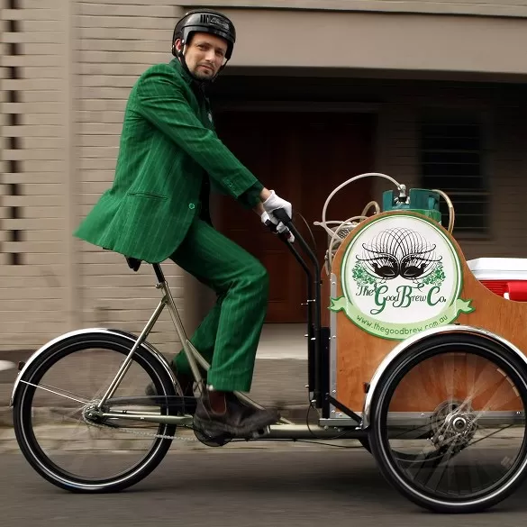 Deano of Good Brew riding a custom bike with a 3D-printed glowing bar top branded with the Good Brew logo, representing the brand's origin as an environmental project delivering kombucha via solar-powered bikes.