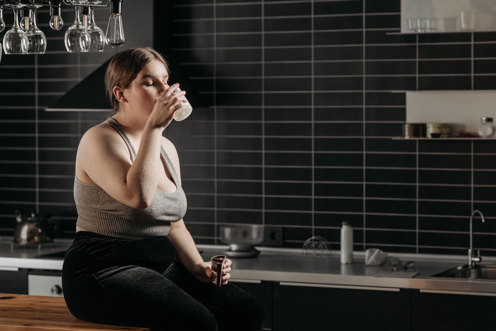 Woman sipping a glass of kombucha at a kitchen counter in the evening, at least one hour before bedtime.