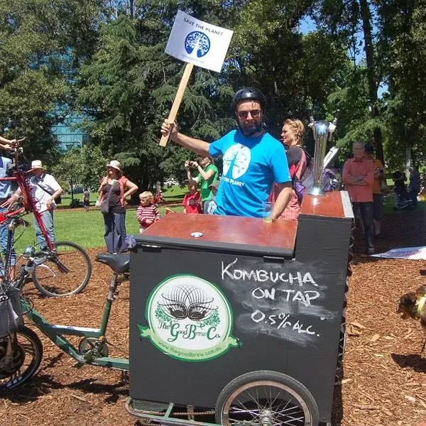 Good Brew kombucha on tap at a community event, with Deano holding a 'Save the Planet' sign near the branded black cart, showcasing live kombucha distribution at festivals.